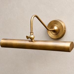 May include: A wall-mounted picture light with a brushed brass finish. The fixture features a cylindrical horizontal shade attached to a curved arm, which connects to a circular wall plate. A small adjustable knob is located at the joint.