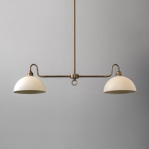 May include: A two-light pendant light fitting with a vintage brass finish. The fixture features two off-white, dome-shaped shades. The light hangs from a central rod with curved arms extending to each shade. A ring detail is centered.