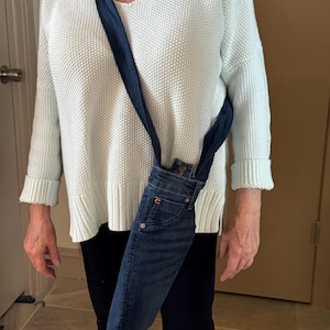 May include: A denim crossbody bag made from repurposed jeans. The bag features the front pockets and waistband of the jeans, with a long strap for over-the-shoulder wear. The denim is a medium wash blue.