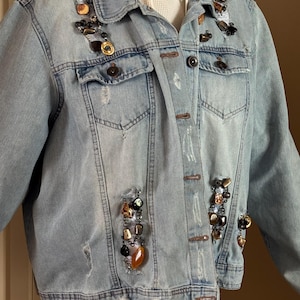 May include: Light blue denim jacket with distressed details and decorative beaded embellishments. The jacket features multiple pockets, button closures, and a classic collar. The embellishments include a mix of black, gold, and brown beads and stones.