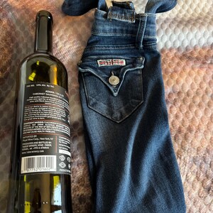 May include: A dark glass wine bottle next to a denim wine bottle bag. The bag is made from a pair of jeans, with the back pocket and button detail visible. The bottle has a black label with white text.