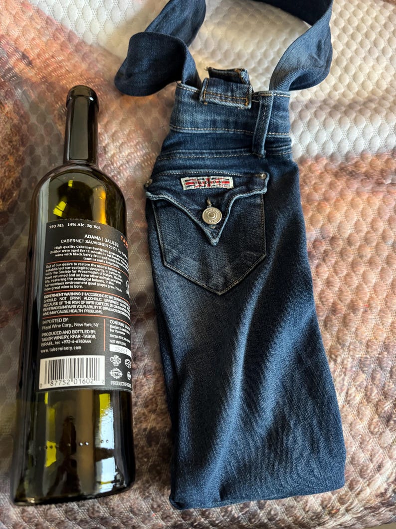 May include: A dark glass wine bottle next to a denim wine bottle bag. The bag is made from a pair of jeans, with the back pocket and button detail visible. The bottle has a black label with white text.