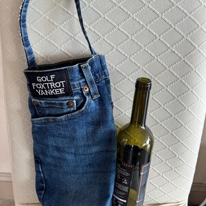 May include: A denim wine bottle carrier with a handle, featuring a pocket and the text "GOLF FOXTROT YANKEE" on a patch. A green wine bottle is placed inside the carrier. The carrier is made from blue jeans material.