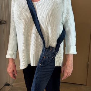 May include: A denim crossbody bag made from recycled jeans. The bag features a dark blue denim exterior with a strap made from the same material. The bag is worn over the shoulder and hangs down the side.