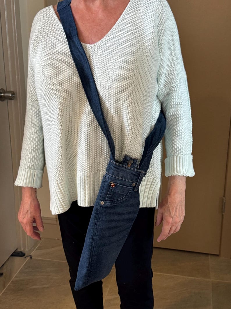 May include: A denim crossbody bag made from recycled jeans. The bag features a dark blue denim exterior with a strap made from the same material. The bag is worn over the shoulder and hangs down the side.