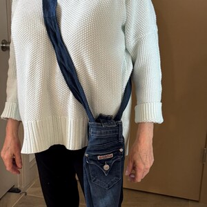 May include: A denim crossbody bag made from repurposed jeans. The bag features the back pocket and button closure of the jeans, with a dark blue strap. The bag is a unique, eco-friendly accessory.
