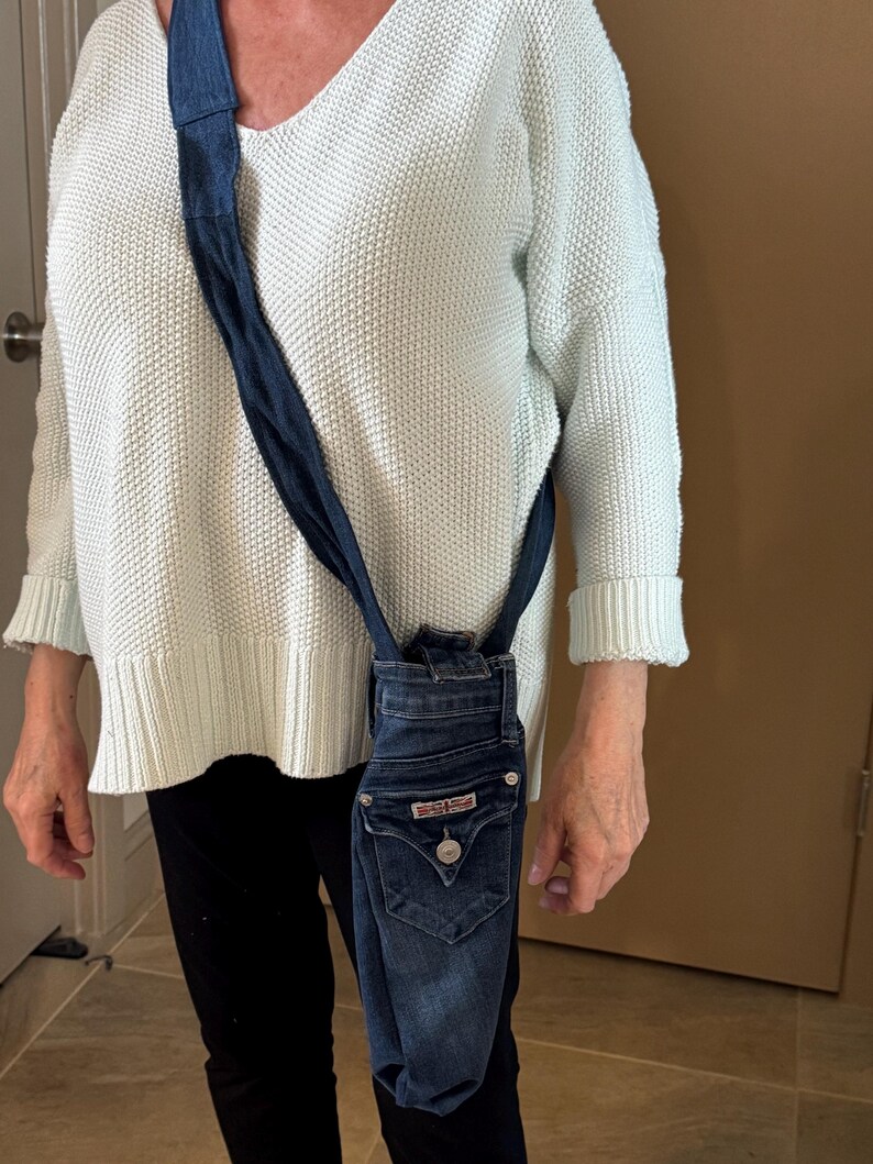 May include: A denim crossbody bag made from repurposed jeans. The bag features the back pocket and button closure of the jeans, with a dark blue strap. The bag is a unique, eco-friendly accessory.