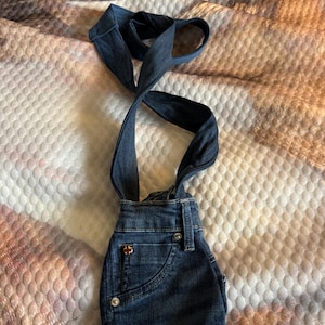 May include: Dark blue denim overalls with straps. The overalls have a small pocket on the front and a red and white logo. The straps are crossed and looped at the top. The overalls are made of a sturdy denim material.