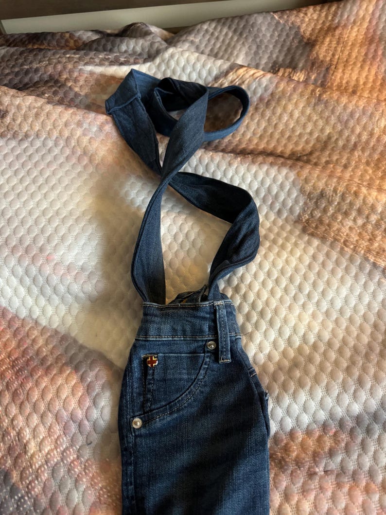 May include: Dark blue denim overalls with straps. The overalls have a small pocket on the front and a red and white logo. The straps are crossed and looped at the top. The overalls are made of a sturdy denim material.
