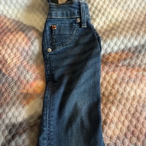 May include: A denim bottle cover shaped like a pair of overalls. The blue jean material has a small pocket with a decorative emblem. The straps extend upwards, creating a unique design.