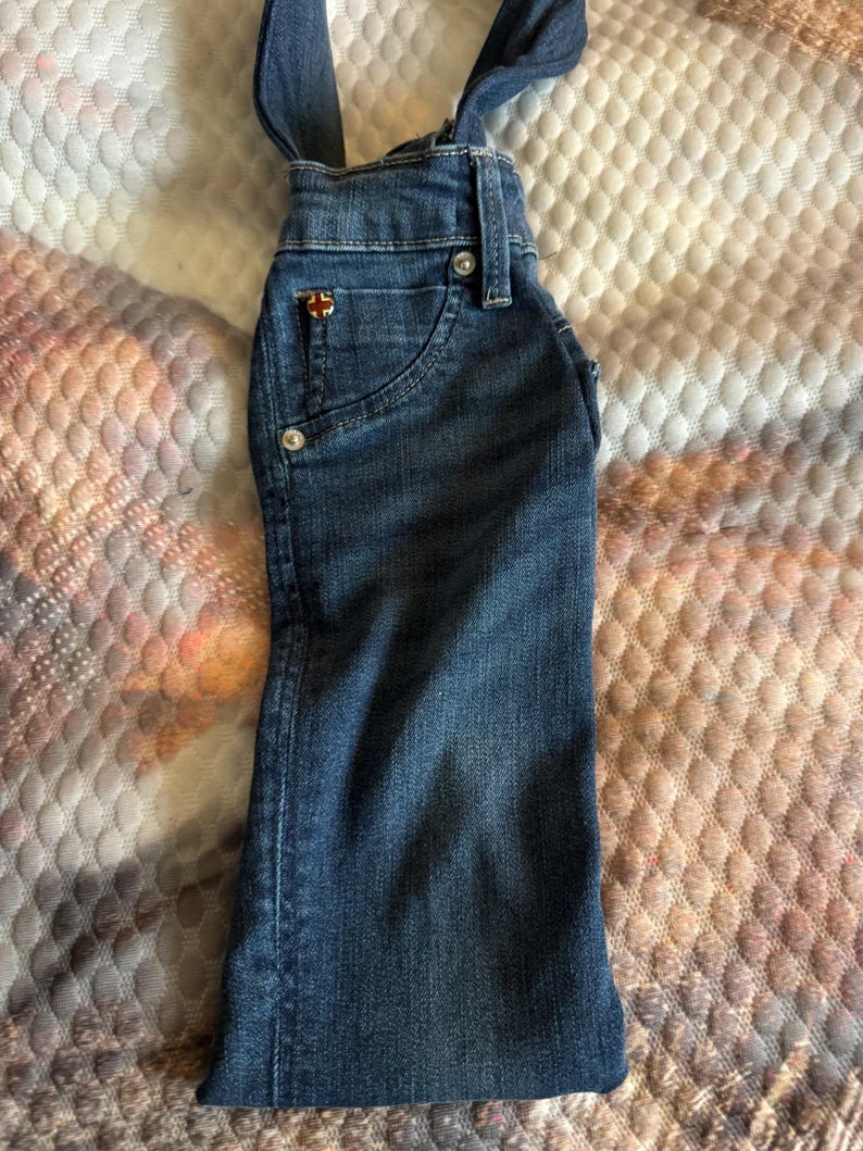 May include: A denim bottle cover shaped like a pair of overalls. The blue jean material has a small pocket with a decorative emblem. The straps extend upwards, creating a unique design.