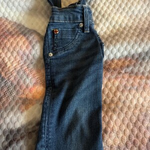 May include: Dark blue denim overalls with adjustable straps. Features a small pocket on the front with a decorative emblem and two front pockets. The overalls have silver-colored button closures and are made of a sturdy denim material.