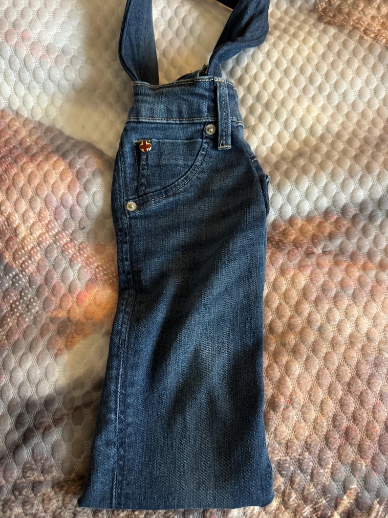 May include: Dark blue denim overalls with adjustable straps. Features a small pocket on the front with a decorative emblem and two front pockets. The overalls have silver-colored button closures and are made of a sturdy denim material.