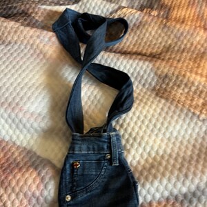 May include: Dark blue denim overalls with suspender straps. The overalls have a front pocket, silver buttons, and a small logo on the front. The suspender straps are crossed and looped at the top.