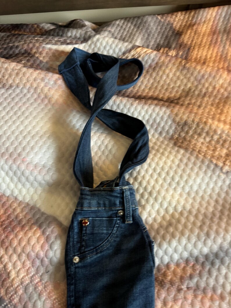 May include: Dark blue denim overalls with suspender straps. The overalls have a front pocket, silver buttons, and a small logo on the front. The suspender straps are crossed and looped at the top.