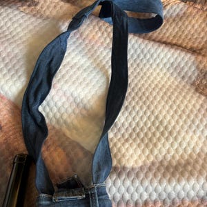 May include: A denim bag with a strap, made from repurposed jeans. The bag features the back pockets and waistband of the jeans, with the straps extending upwards. The denim is a medium blue wash.