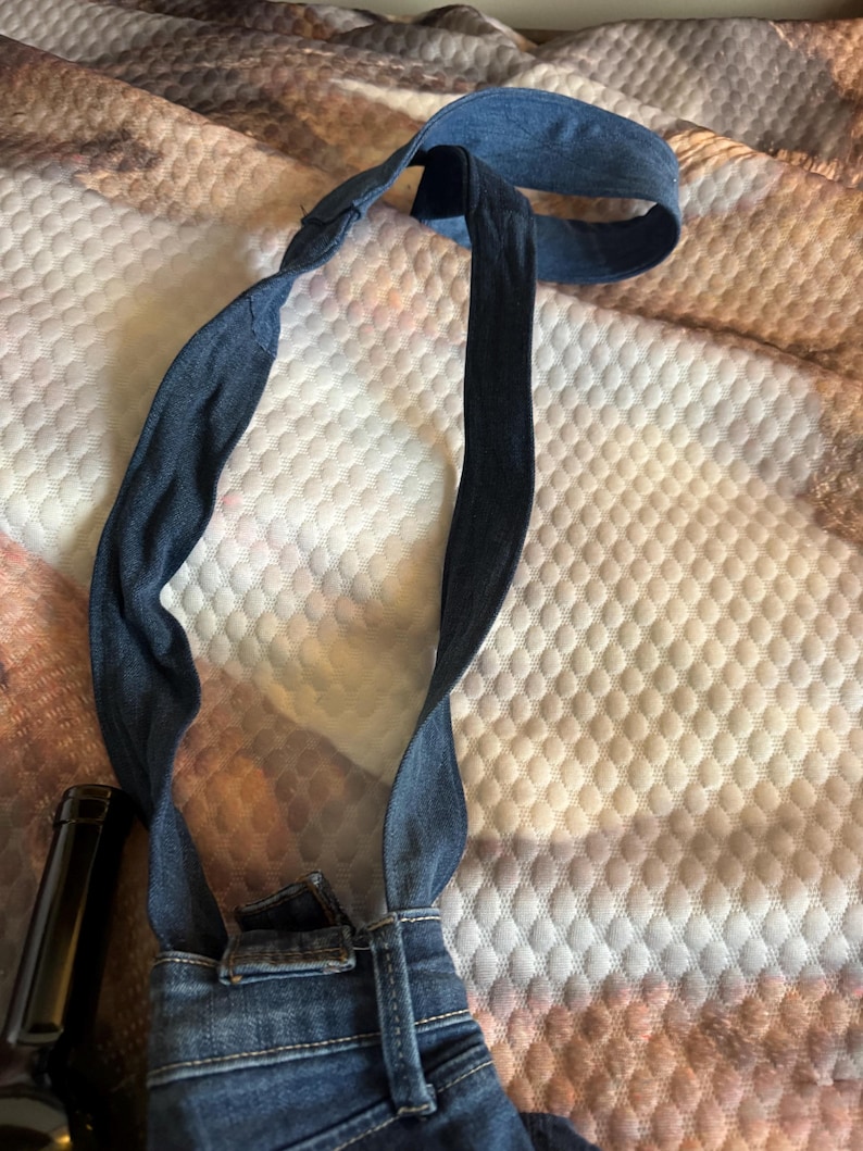 May include: A denim bag with a strap, made from repurposed jeans. The bag features the back pockets and waistband of the jeans, with the straps extending upwards. The denim is a medium blue wash.
