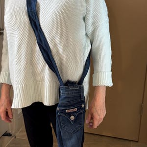 May include: A denim crossbody bag made from repurposed jeans. The bag features the back pockets and waistband of the jeans, with a dark blue strap. The bag is a unique, eco-friendly accessory.
