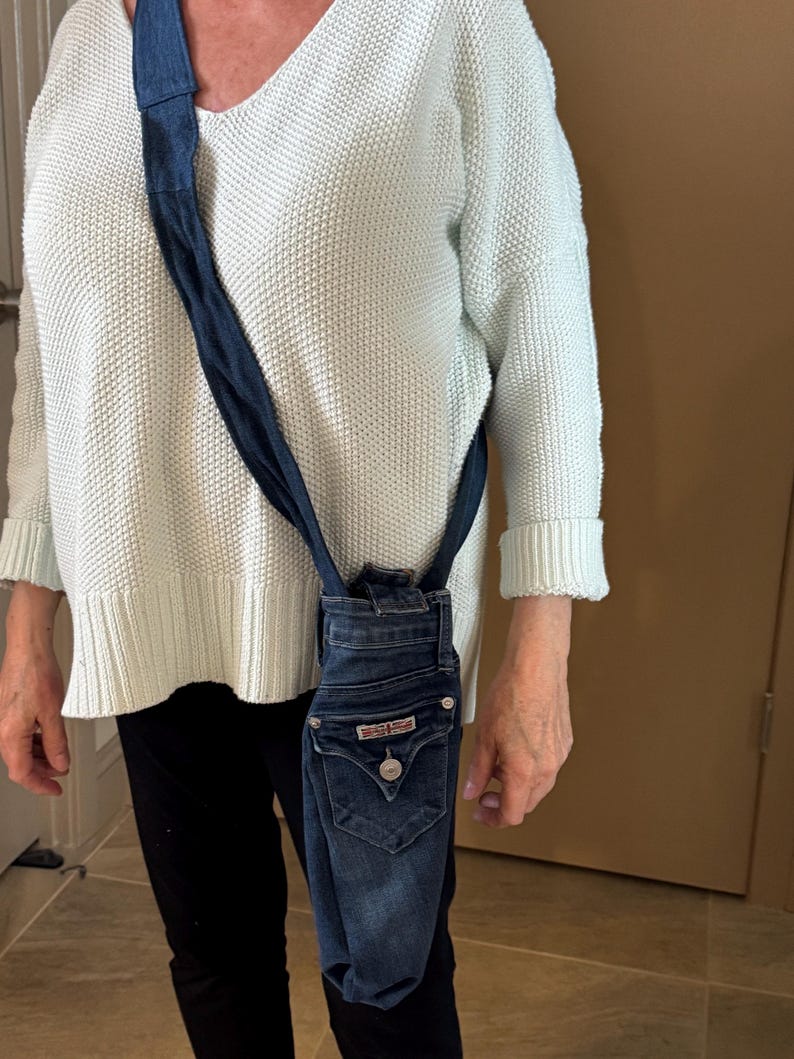 May include: A denim crossbody bag made from repurposed jeans. The bag features the back pockets and waistband of the jeans, with a dark blue strap. The bag is a unique, eco-friendly accessory.