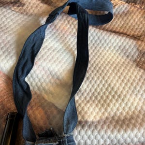 May include: A denim bib overall strap, featuring a dark blue color. The strap is connected to the waistband of a pair of denim overalls. The strap is looped and appears to be adjustable.