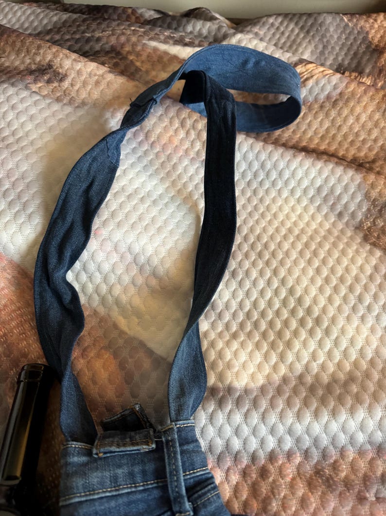 May include: A denim bib overall strap, featuring a dark blue color. The strap is connected to the waistband of a pair of denim overalls. The strap is looped and appears to be adjustable.