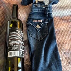 May include: A dark glass wine bottle next to a denim wine bottle bag. The bottle has a black label with white text. The denim bag is made from blue jeans with a pocket and a button.