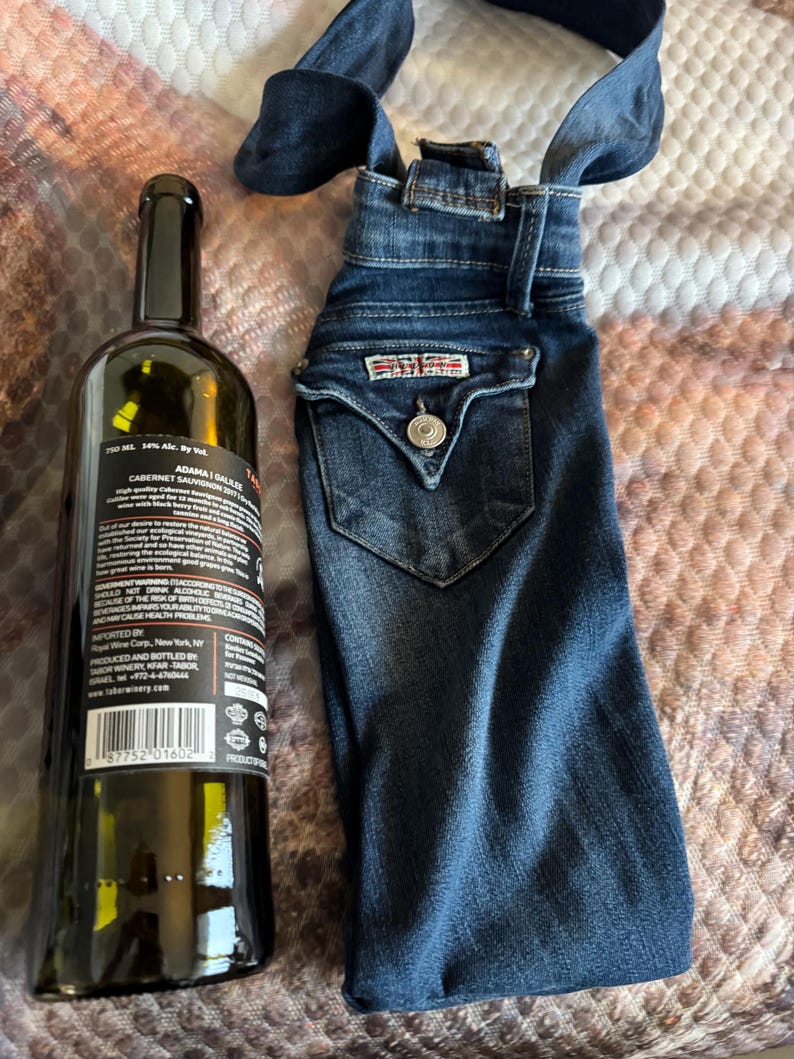 May include: A dark glass wine bottle next to a denim wine bottle bag. The bottle has a black label with white text. The denim bag is made from blue jeans with a pocket and a button.