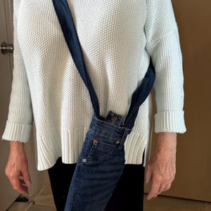 May include: A denim crossbody bag made from repurposed jeans. The bag features the front pocket and waistband of the jeans, with a dark blue strap. The bag is worn over the shoulder.