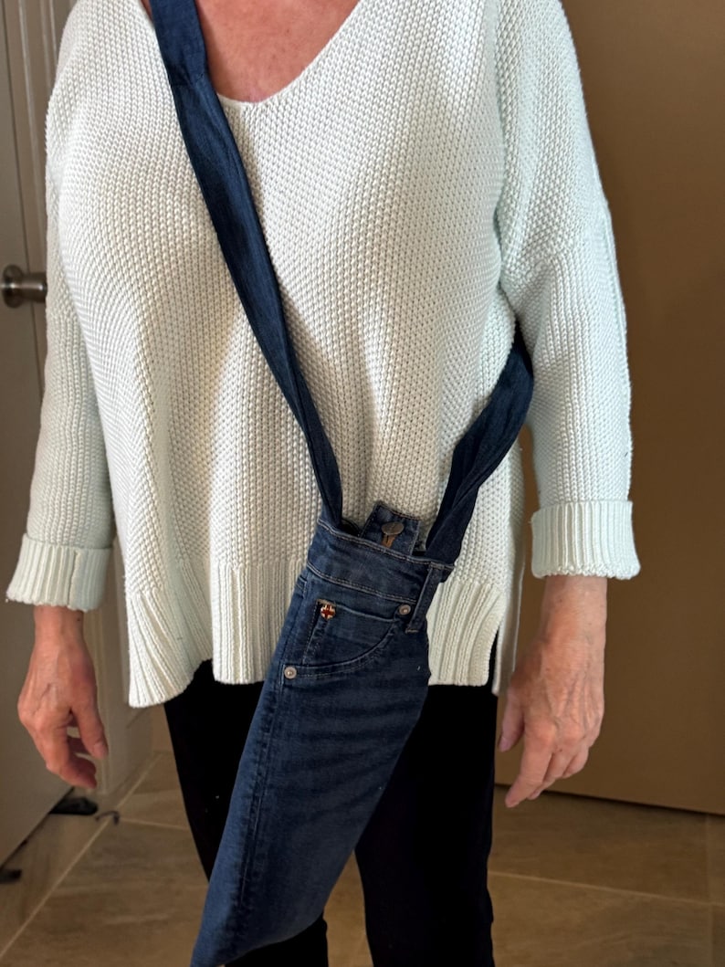May include: A denim crossbody bag made from repurposed jeans. The bag features the front pocket and waistband of the jeans, with a dark blue strap. The bag is worn over the shoulder.