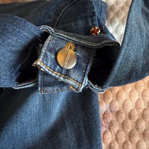 May include: Close-up of a blue denim garment, possibly a jacket or shirt, with visible stitching and metal buttons. One button is round with the brand name "Carhartt" engraved. Another button has a red cross design.