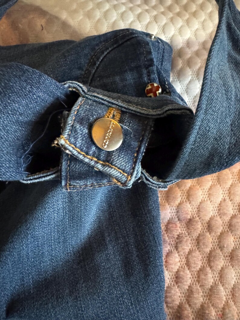 May include: Close-up of a blue denim garment, possibly a jacket or shirt, with visible stitching and metal buttons. One button is round with the brand name "Carhartt" engraved. Another button has a red cross design.