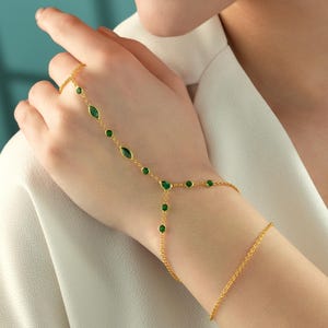 May include: A gold hand chain bracelet featuring a series of small round and marquise-cut green stones. The jewelry piece connects a ring component worn on the finger to a delicate chain that wraps around the wrist with a secure clasp closure.