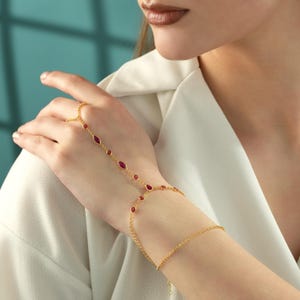 May include: A gold hand chain bracelet featuring a delicate chain that connects a ring to a wrist bracelet. The jewellery is adorned with small, red, marquise-cut and round-cut gemstones set in gold bezels, creating a continuous decorative link.