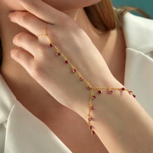 May include: A gold hand chain bracelet featuring a delicate chain that connects a ring to a wrist bracelet. The jewellery is adorned with small, round, bezel-set magenta-pink stones spaced evenly along the entire length of the gold-toned metal chain.