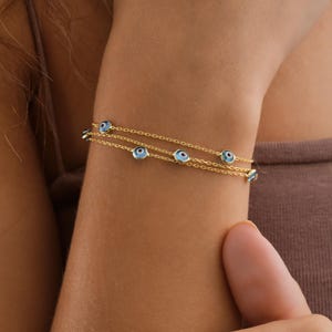 Könnte beinhalten: Ein zartes Goldkettenarmband mit mehreren kleinen, kreisförmigen Nazar-Amuletten. Jeder Anhänger besteht aus einer hellblauen Glasperle mit einem dunkelblauen und weißen Zentrum, die gleichmäßig entlang der dünnen, polierten Goldkettenelemente verteilt sind.