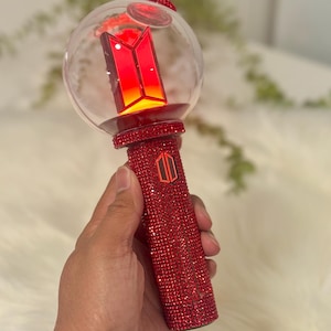 May include: A red, bedazzled light stick with a clear, spherical top. The top features a red logo and illuminates with a warm glow. The handle is covered in red rhinestones, and the overall design is eye-catching.