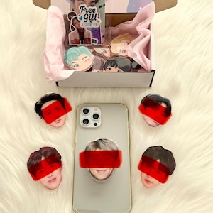 May include: A clear phone case with a pop-up grip featuring a face with a red bar over the eyes, surrounded by similar face-shaped stickers. A box with stickers and a "Free Gift" card is also visible.