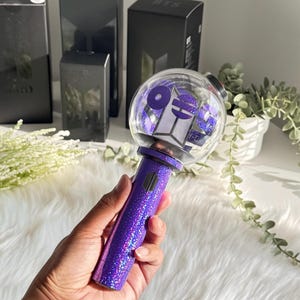 May include: A purple glitter light stick with a clear orb containing purple confetti and a logo. The light stick is held in a hand, with several black boxes in the background. The boxes have the letters "BTS" printed on them.
