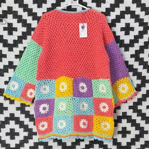 May include: A colorful, handmade crochet cardigan. The top is coral, with mint green and purple sleeves. The bottom features daisy squares in various colors. The cardigan is hanging on a white hanger against a black and white patterned background.