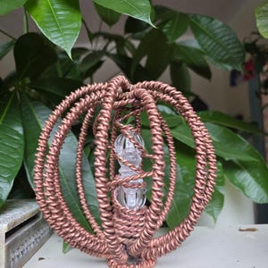 May include: A copper wire sculpture with a clear glass element at its core. The spherical sculpture has concentric rings of twisted copper wire. Green leaves form the background.