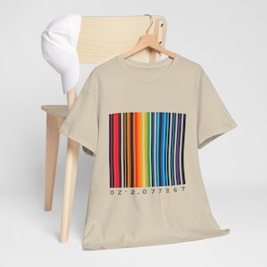Pode incluir: Uma camiseta bege com um design de código de barras colorido. O código de barras apresenta listras verticais em várias cores, incluindo vermelho, laranja, amarelo, verde, azul e roxo. Abaixo do código de barras está a sequência numérica "02.2.077967". Um boné de beisebol branco repousa sobre uma cadeira de madeira.