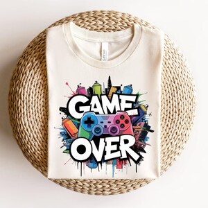 May include: Cream-colored t-shirt with a colorful graphic design. The design features a video game controller, spray paint cans, and the words "GAME OVER" in bold, white letters. The shirt is laid on a woven, circular surface.