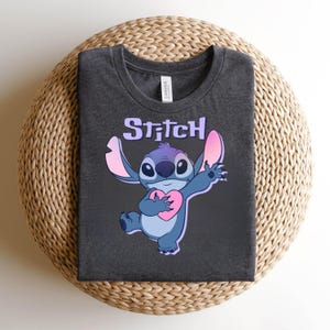 May include: Dark gray t-shirt featuring a cartoon character, Stitch, holding a pink heart. The word "Stitch" is printed above the character in a light purple color. The shirt is folded on a woven surface.
