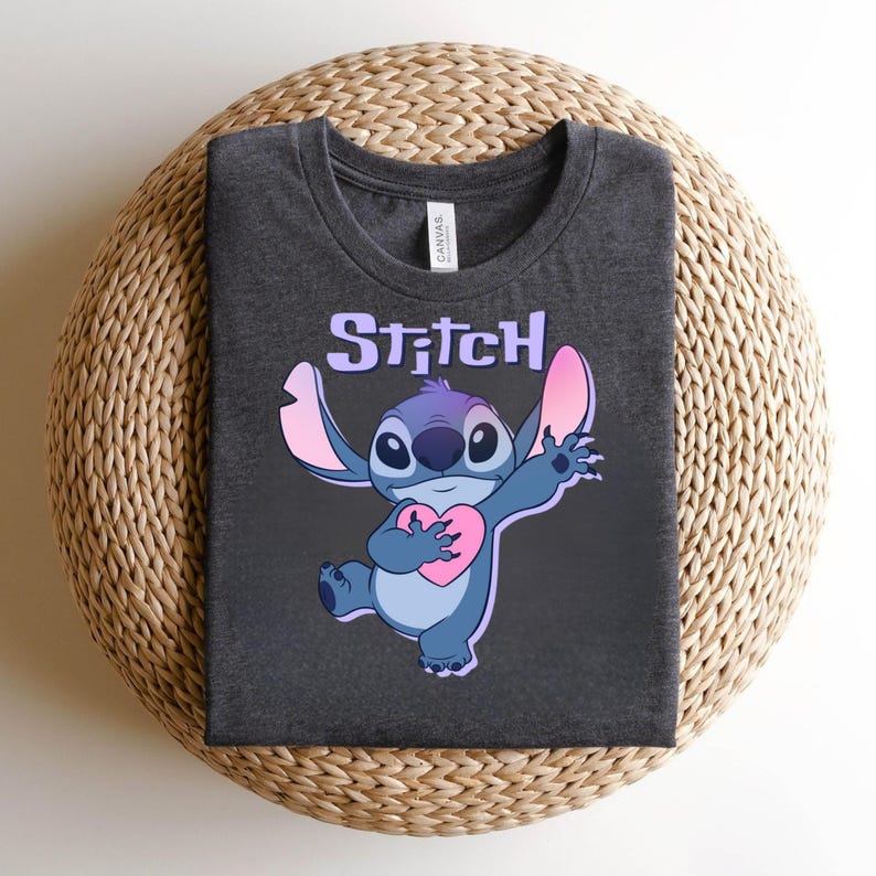 May include: Dark gray t-shirt featuring a cartoon character, Stitch, holding a pink heart. The word "Stitch" is printed above the character in a light purple color. The shirt is folded on a woven surface.