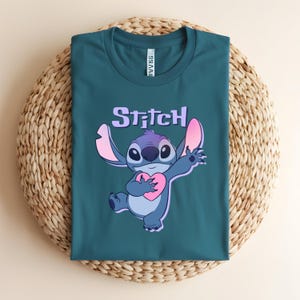 May include: Teal t-shirt featuring a cartoon image of Stitch holding a pink heart. The word "Stitch" is printed above the character in a purple, bubble-style font. The shirt is folded on a woven, circular surface.