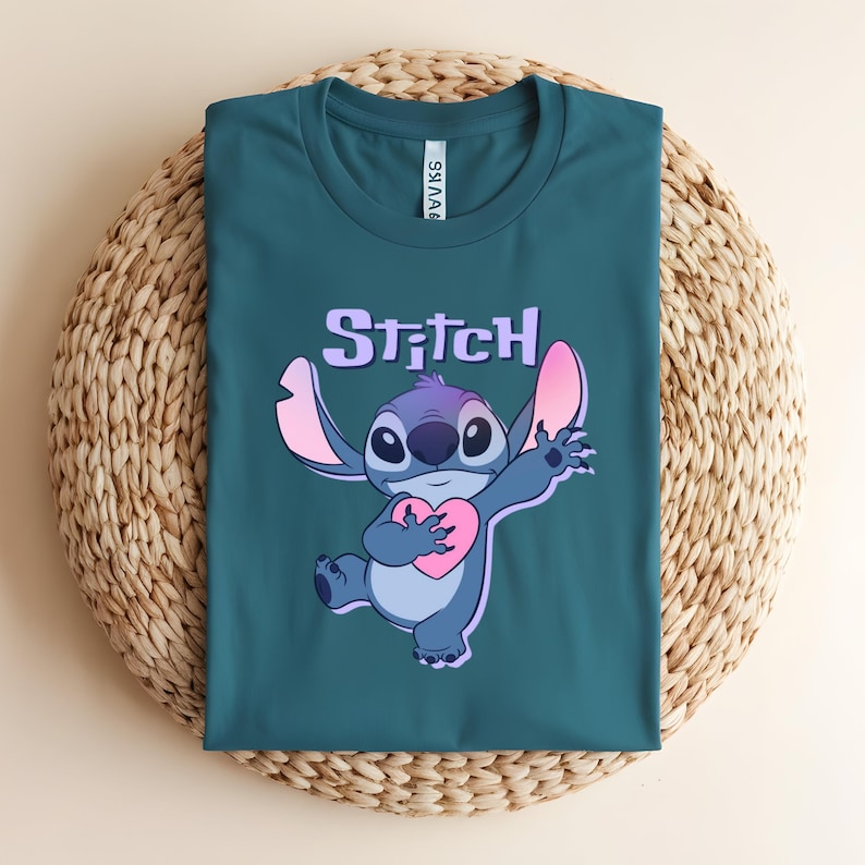 May include: Teal t-shirt featuring a cartoon image of Stitch holding a pink heart. The word "Stitch" is printed above the character in a purple, bubble-style font. The shirt is folded on a woven, circular surface.