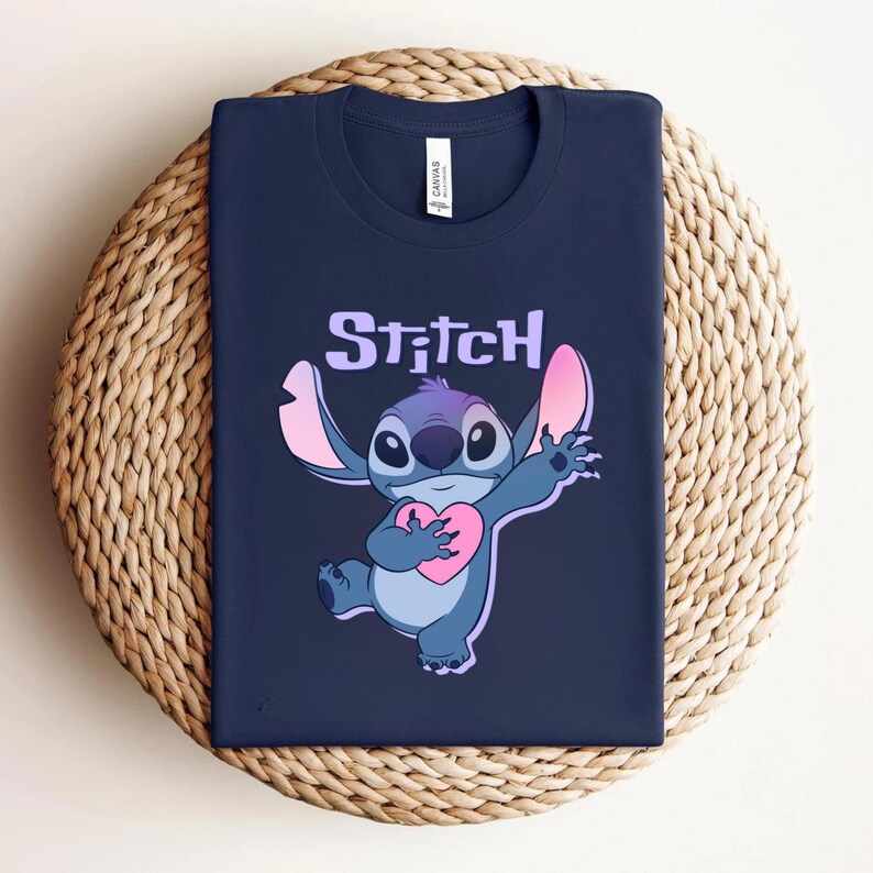 May include: Navy blue t-shirt featuring a cartoon image of Stitch holding a pink heart. The word "Stitch" is printed above the character in a purple font. The shirt is folded and placed on a woven, circular surface.