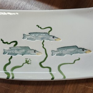 May include: White rectangular ceramic serving dish with rounded corners. The dish is decorated with three stylized gray fish with yellow beaks and green seaweed. The dish is likely used for serving sushi or appetizers.