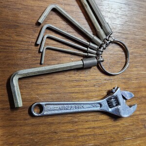 May include: A set of five L-shaped hex keys of varying sizes attached to a circular metal ring, and a single silver-colored adjustable wrench with the brand name Craftsman embossed on the handle, both made of metal with a matte finish.