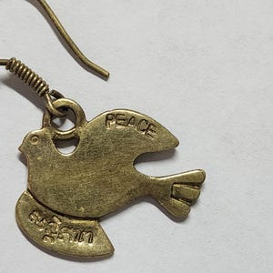 May include: A single brass-colored earring featuring a dove design. The word "PEACE" is inscribed on the wing, and other text is visible on the lower portion of the bird. The earring has a hook closure.